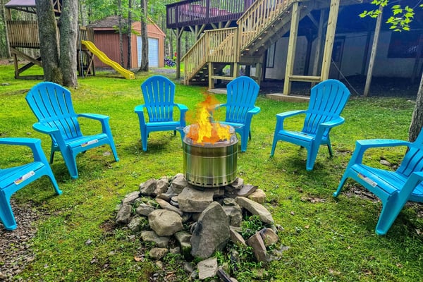 Backyard fire pit surrounded by bright blue Adirondack chairs, with cabin and elevated deck in wooded setting