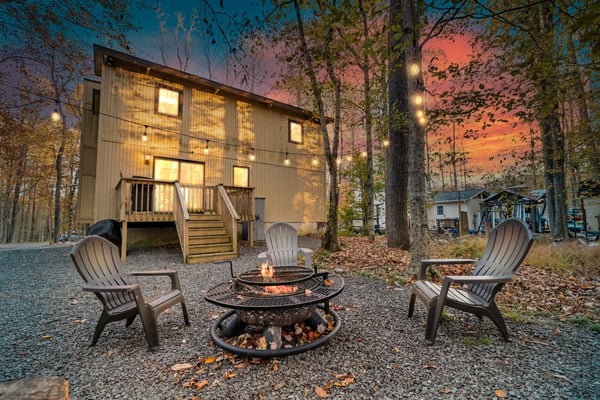 Modern cabin with lit windows surrounded by tall trees, fire pit with two Adirondack chairs in foreground at dusk.