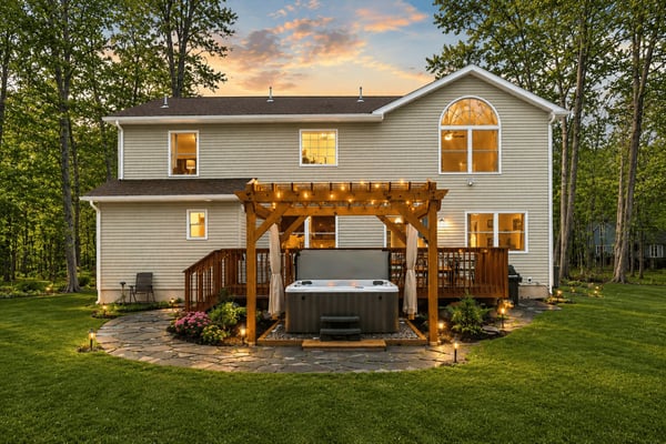 Two-story house with hot tub under wooden pergola, lit deck, and surrounding trees at dusk
