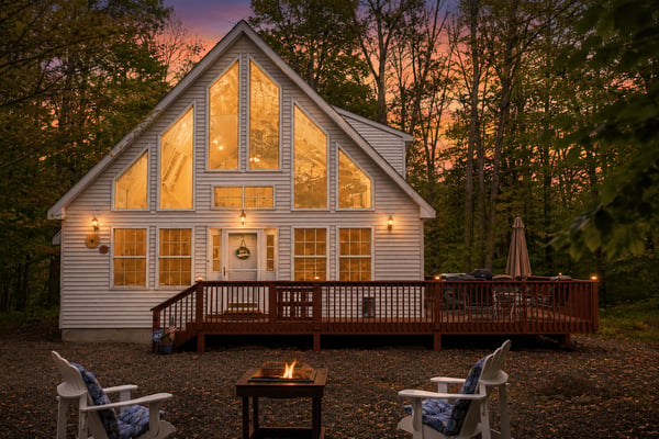 Modern white cottage with large windows glowing at dusk, surrounded by trees, with fire pit and Adirondack chairs in foreground.