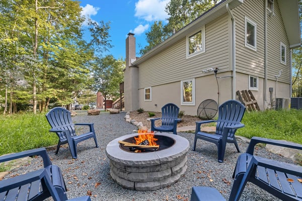 Backyard fire pit with blue Adirondack chairs, stone surround, and two-story house with siding surrounded by trees