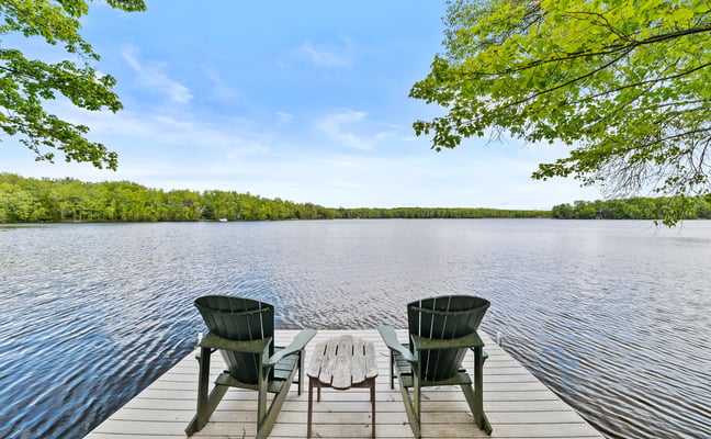 Two green Adirondack chairs on a wooden dock facing a calm lake with forested shoreline and blue sky